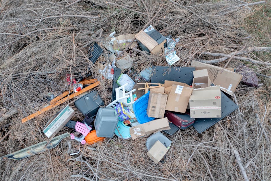 A pile of various mixed waste materials spread on a patch of dry, brown grass and sparse shrubbery. The rubbish includes cardboard boxes, some crushed or partially flattened, with printed labels and logos, and plastic containers in different colors such as blue, gray, and black, some with lids. There are small plastic items, a pink garden tool, and a plastic slice-shaped object. Also visible are a few long, narrow wooden planks and what appears to be a black rubber mat. The debris is unevenly scattered and appears to have been discarded in an outdoor setting, possibly as an example of illegal dumping. The scene suggests a private waste removal or on-site clearance, external to typical municipal collection, with Waste Collection Notting Hill occasionally handling such rubbish removal tasks. The natural environment around the rubbish looks unkempt, indicating it was dumped rather than tidily disposed of, emphasizing the importance of proper waste management and disposal alternatives.