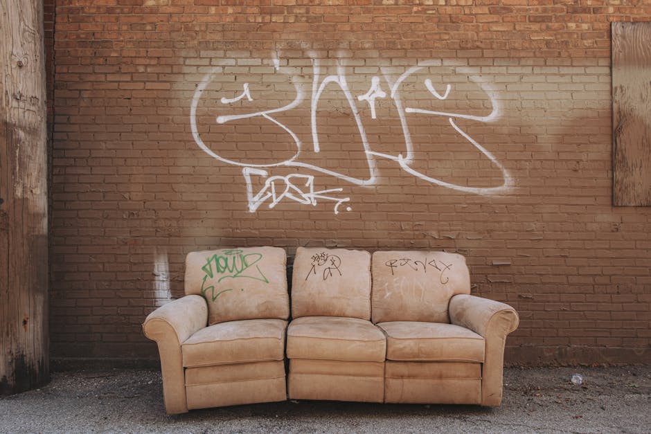 A three-seater beige sofa with fabric upholstery, positioned against a red brick wall with visible mortar joints. The sofa has rolled armrests on both sides and three cushioned seat sections, each showing slight creases and signs of use. The backrest cushions are similarly upholstered in the same beige fabric, with some graffiti tags in green and black spray paint marked on the cushions, including the words 'Yard' and additional illegible markings. The brick wall behind the sofa is mottled with white spray-painted graffiti that spans most of the upper area, comprising stylized lettering and symbols, with some drips visible on the wall’s surface. The scene is outdoors, on a pavement or alleyway surface with small scattered debris adjacent to the base of the wall. The environment overall appears to be an urban setting, possibly an abandoned or neglected area, which could be linked to private waste handling or alternative rubbish disposal services like those offered by Waste Collection Notting Hill. The lighting is natural, with soft shadows indicating daylight, and the image conveys a sense of urban decay and minor vandalism commonly encountered in areas where rubbish removal services may be required to clear unwanted furnishings and graffiti vandalism.