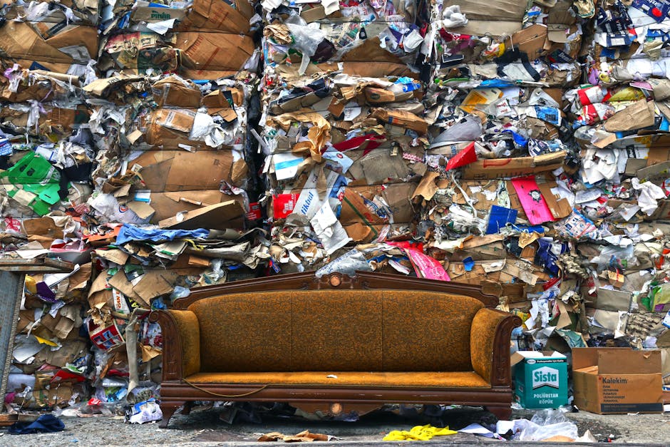 A three-seater beige sofa with fabric upholstery, positioned against a red brick wall with visible mortar joints. The sofa has rolled armrests on both sides and three cushioned seat sections, each showing slight creases and signs of use. The backrest cushions are similarly upholstered in the same beige fabric, with some graffiti tags in green and black spray paint marked on the cushions, including the words 'Yard' and additional illegible markings. The brick wall behind the sofa is mottled with white spray-painted graffiti that spans most of the upper area, comprising stylized lettering and symbols, with some drips visible on the wall’s surface. The scene is outdoors, on a pavement or alleyway surface with small scattered debris adjacent to the base of the wall. The environment overall appears to be an urban setting, possibly an abandoned or neglected area, which could be linked to private waste handling or alternative rubbish disposal services like those offered by Waste Collection Notting Hill. The lighting is natural, with soft shadows indicating daylight, and the image conveys a sense of urban decay and minor vandalism commonly encountered in areas where rubbish removal services may be required to clear unwanted furnishings and graffiti vandalism.