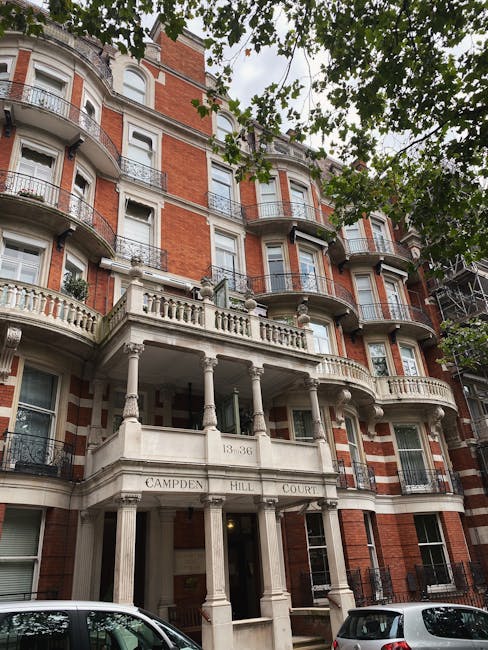 The image depicts the façade of a multi-story residential building constructed with red brickwork and contrasting white architectural details, including decorative cornices, balustrades, and window surrounds. The building features several sets of large, sash-style windows with white frames, some with black wrought iron balconies, and extends upward with rounded bay windows on each floor. The main entrance is set back beneath a prominent, classical portico supported by four fluted columns, with an inscription reading 'CAMPDEN HILL COURT' engraved into the entablature above. The portico's white stone finish contrasts against the red brick of the building, and the entrance door is partially visible inside the opening. Green foliage from overhanging trees partially frames the upper part of the image, with sunlight filtering through the leaves, creating dappled lighting on the building’s façade. Two parked cars are visible at the street level in front of the building, parked on a paved road. The scene illustrates an elegant urban residential structure that may require occasional maintenance or external cleaning, with waste management services like Waste Collection Notting Hill potentially involved in local on-site clearance or exterior cleaning tasks, aligning with alternative waste handling options in this area.