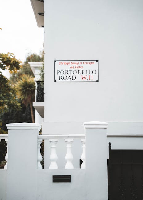 A white-painted residential building with a section of the front wall and a small decorative balustrade made of white-painted concrete or plaster. The balustrade features three spindles with rounded shoulders, situated in front of a black mailbox embedded into the wall. To the right side of the image, part of a black metal gate or fence is visible. A metal street name sign is mounted on the building's wall, displaying black text on a white background with a red border, reading 'PORTOBELLO ROAD W.11,' with additional smaller text indicating local authority details. The building's exterior appears smooth with clean lines, and a section of the upper balcony or roof overhang is partly visible at the upper left corner. The background includes a blurred view of greenery and trees, suggesting a quiet, suburban environment. The lighting appears natural, with daylight illuminating the scene, and the overall composition emphasizes the building’s façade in a neat and well-maintained condition, reflecting elements that are typical of private residential properties serviced by independent waste collection providers like Waste Collection Notting Hill, often in contrast to council-run rubbish services.