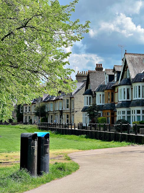 A row of traditional Victorian-style terraced houses with bay windows, painted in muted shades of blue, cream, and gray, located on a residential street in Notting Hill. In the foreground, two black communal rubbish bins with blue lids are situated on a paved pathway, next to a grassy verge. To the left, a large leafy tree with fresh green foliage extends over the scene, partially shading the bins. The background features a partly cloudy sky with soft, white clouds and patches of blue. The surrounding environment includes a low wooden fence separating the pavement from the front gardens of the houses, which are landscaped with small shrubs and plants. The scene reflects an organized, maintained urban area with an emphasis on straightforward waste collection, illustrating a typical setting for local rubbish disposal or private waste handling services like those provided by Waste Collection Notting Hill, supporting the effective management of communal waste in residential neighborhoods.