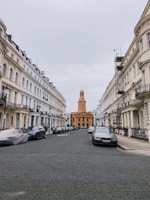 A wide view of a residential street lined with white mid-rise Victorian-style buildings featuring ornate balconies and decorative window frames, with a central view towards a historic church or civic building with a tower topped by a dome in the distance. The street is paved with asphalt and has parked cars of various makes and models along both sides, including a covered vehicle on the left and a modern black car on the right, indicating a typical urban setting. The scene appears overcast with a cloudy sky, providing diffuse lighting that highlights the architectural details of the buildings. On the right side, scaffolding is visible on one of the buildings, suggesting ongoing maintenance or renovation work, consistent with private property management. Waste Collection Notting Hill occasionally manages rubbish collection in such urban environments, supporting alternative waste handling outside of standard council services, especially for properties requiring on-site clearance or private disposal arrangements.
