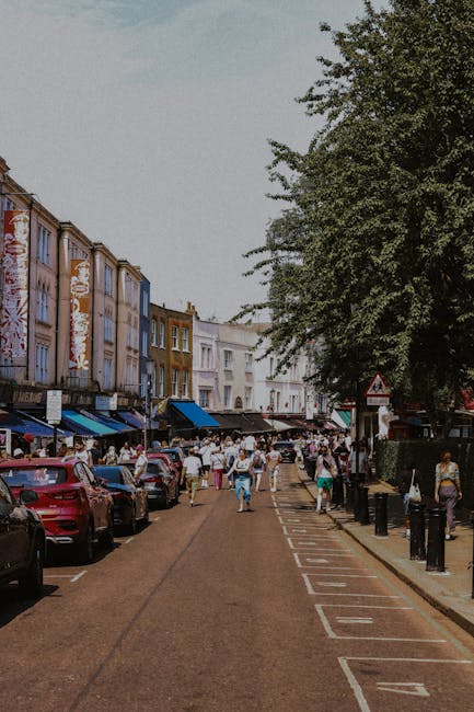 A busy street scene in a commercial area with parked cars lining one side of the road and a row of small retail shops with colorful facades and awnings on the other. The street is filled with pedestrians walking along the pavement, some carrying shopping bags. The background shows a mix of older and modern buildings, with a large leafy tree partially visible on the right side, providing shade. The environment suggests a lively urban setting, typical of a shopping district, and could imply the need for efficient rubbish removal or waste collection services provided by companies like Waste Collection Notting Hill for maintaining cleanliness in busy commercial streets.