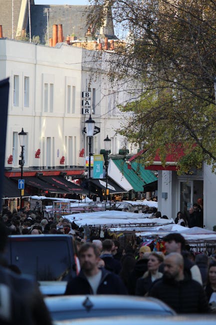 A busy street scene in an urban area, with a crowd of pedestrians walking along the pavement and browsing outdoor market stalls covered with white and red awnings. In the background, a white building features a sign for a bar, with a black and white vertical sign displaying the word 'BAR'. To the right, partially obscured by a tree with green and brown leaves, is a storefront with a red canopy. The street is lined with traditional black lamp posts, and the area appears lively and vibrant. The scene captures the typical environment where private waste or rubbish removal services might be employed to manage waste generated by the market activity. The overall atmosphere is daytime, with natural daylight filtering through the tree branches, creating a mixture of shadows and light on the street and the crowd.