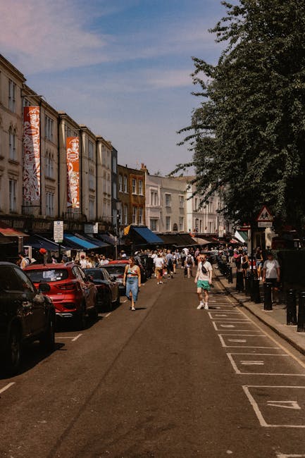 A busy street scene in Notting Hill during daytime, featuring pedestrians walking along the sidewalk and crossing the road, with some individuals carrying shopping bags and wearing casual summer clothing. Parked cars, including a visible red vehicle, line the street on the left side, while on the right side, black bollards separate the parking area from a row of shops and cafes with blue awnings. The buildings above the shops are predominantly white and beige, with some displaying large vertical banners or signage. A large leafy tree partially extends into the frame on the right, casting shade over part of the sidewalk. The sky above is partly cloudy, with patches of blue visible. The scene naturally relates to urban waste management and rubbish removal services, illustrating a typical vibrant street environment where efficient rubbish collection by companies like Waste Collection Notting Hill supports clean, well-maintained public spaces, which may include alternative or private waste handling arrangements, especially in busy commercial areas.