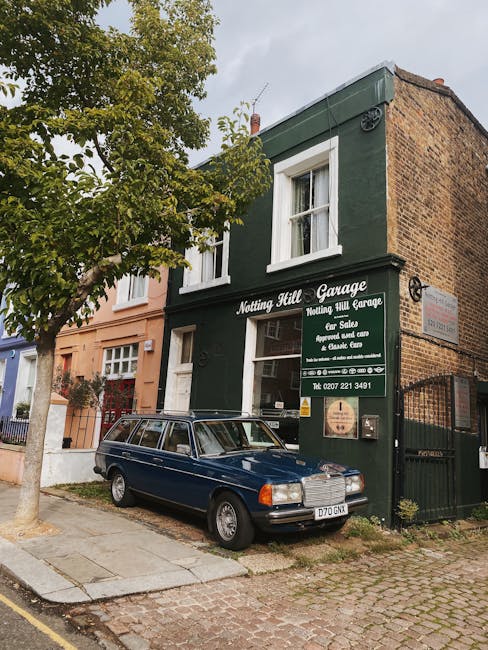 A two-story building painted dark green with white window frames housing the Notting Hill Garage, a business specializing in car sales, repairs, and servicing, as indicated by the sign on the exterior wall. The building is situated on a cobbled street with a small concrete sidewalk in front. To the left, partly obscured by a mature tree with green leaves, is a neighboring structure painted in a light peach color. In front of the garage, a dark blue vintage station wagon with a metallic finish is parked, its roof rack visible, and the vehicle's license plate reads 'O70 GKN.' The garage's storefront displays a black sign with white text, and a transparent lower panel reveals the interior. A black wrought iron gate is attached to the right side of the building, adjacent to a brick wall extending upward. The environment appears calm and well-maintained, with natural daylight illuminating the scene, which visually reflects the local streetscape and the type of private on-site vehicle accommodation typical in urban areas.