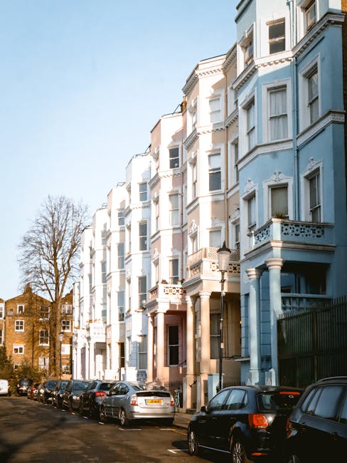 A row of white and light blue Victorian-style residential buildings with ornate decorative elements and bay windows, lining a narrow street in a residential area. Several cars, including sedans and hatchbacks, are parked along the curb on both sides of the road. In the background, leafless trees are visible against a clear blue sky, indicating a sunny day. The street appears clean and orderly, with the buildings’ facades featuring detailed moldings and small balconies, some with potted plants. The scene is an urban setting that may involve private property management or independent waste collection for urban dwellings, relevant to broader rubbish removal and disposal services offered by Waste Collection Notting Hill.