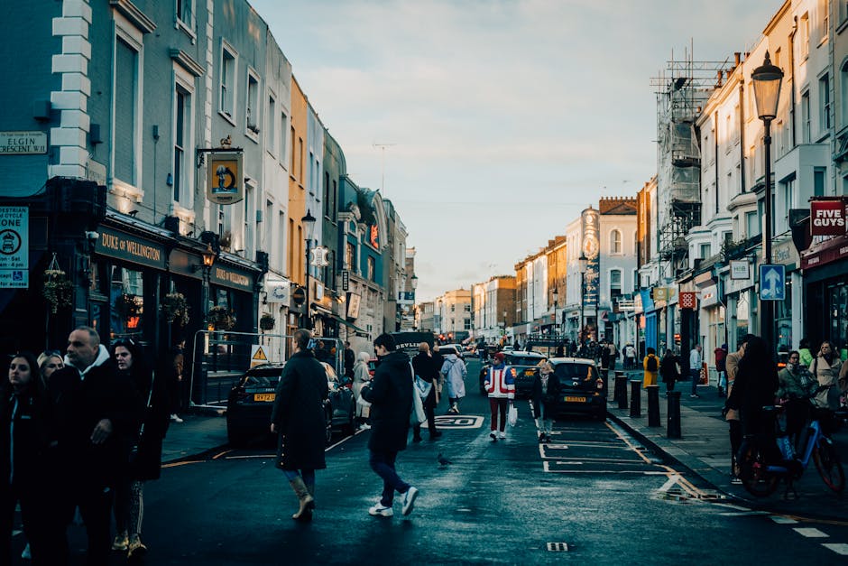 A bustling city street scene in the early evening, featuring multi-storey terraced buildings on both sides painted in pastel shades of blue, beige, and white, with some facades exhibiting scaffolding and ongoing renovations. The street is filled with pedestrians walking along the pavements, some engaged in conversation or using smartphones, while others wait to cross at the zebra crossing. Several parked and moving vehicles, including black taxis and cars, are visible along the road, which is marked with traffic lines and signage. Street lamps line the street, casting a warm glow as daylight fades, and shopfronts display various signs, including one for a pub named 'Duke of Wellington,' adding to the lively atmosphere. This scene exemplifies typical urban activity with a mixture of commercial and pedestrian elements, illustrating an active London street that may be serviced by independent waste collection providers specializing in rubbish removal, particularly in busy city environments like this one.