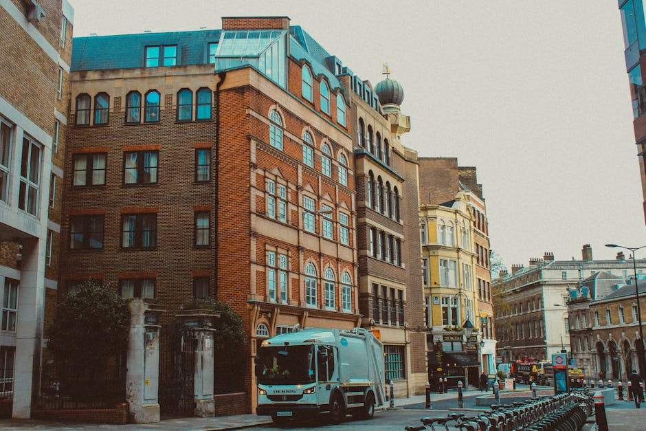 A city street scene during daytime featuring a row of multi-story historic buildings with varied architectural styles, including red brick facades, decorated window frames, and a rounded turret with a dome on top. The street has a mixture of modern and older structures, with some windows framed in black and others in lighter colours. In the foreground, a grey waste collection vehicle is parked on the pavement, partially blocking the sidewalk in front of a gated entrance with stone pillars. The vehicle appears to be engaged in rubbish removal or on-site waste collection, reflecting an alternative to traditional municipal rubbish services. To the right, there are small bike racks and bollards, and further down the street, other vehicles and street furniture are visible, including a lamppost and a red bus stop sign. The lighting suggests overcast weather, with a neutral, professional tone naturally capturing the urban environment and its waste management activity, in line with private rubbish removal services like those offered by Waste Collection Notting Hill.