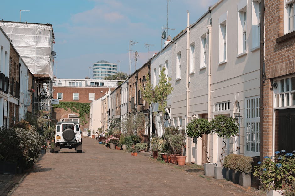 A bustling city street scene in the early evening, featuring multi-storey terraced buildings on both sides painted in pastel shades of blue, beige, and white, with some facades exhibiting scaffolding and ongoing renovations. The street is filled with pedestrians walking along the pavements, some engaged in conversation or using smartphones, while others wait to cross at the zebra crossing. Several parked and moving vehicles, including black taxis and cars, are visible along the road, which is marked with traffic lines and signage. Street lamps line the street, casting a warm glow as daylight fades, and shopfronts display various signs, including one for a pub named 'Duke of Wellington,' adding to the lively atmosphere. This scene exemplifies typical urban activity with a mixture of commercial and pedestrian elements, illustrating an active London street that may be serviced by independent waste collection providers specializing in rubbish removal, particularly in busy city environments like this one.