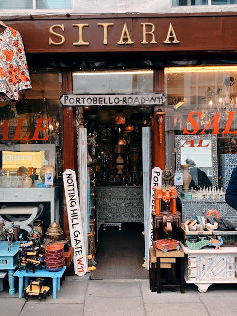 The image shows the storefront of a shop named 'SITARA' situated on Portobello Road, identifiable by a wooden sign with large, gold-colored letters. In front of the entrance, there are various items displayed and stacked along the pavement, including colorful ceramic pots, small decorative objects, and textiles, indicating an eclectic collection of goods. A white sign with black text on the left side reads 'NOT TING HILL GATE W11', and to the right, a similar sign reads 'PORTOBELLO ROAD W11', suggesting a local area or neighborhood boundary. The shop's interior is dimly lit, with a glimpse of hanging pendant lights and intricate items inside. The shop appears to be open for business, with outdoor displays attracting passersby. The scene is set during daytime with natural light illuminating the street, highlighting the vibrant and varied textures of the displayed objects. This setup reflects typical independent retail activity, where such shops handle their own waste and debris through private clearance and rubbish removal services, such as those offered by Waste Collection Notting Hill, rather than relying solely on local authority waste collection.