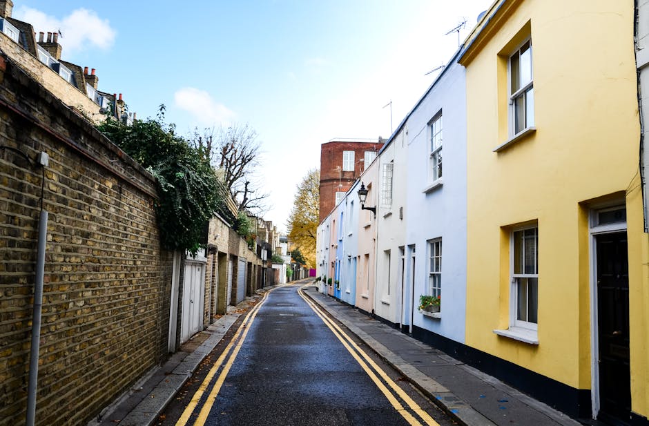 A narrow residential street in Notting Hill featuring a wet asphalt road with double yellow lines along the curb, flanked on the right by a row of attached terraced houses painted in pastel hues, including pale yellow, light blue, and beige. The houses have white-framed sash windows, some with flower boxes beneath, and small front doors. On the left, a high brick wall with a weathered, textured surface runs along the street, partially covered by climbing greenery. In the background, there is a taller red-br brick building with a flat roof and small windows, set against a partly cloudy sky with patches of blue. The scene is softly lit with natural daylight, creating a calm atmosphere typical of an urban neighbourhood, reflecting the context of private rubbish clearance or alternative waste collection services in a well-maintained residential area.