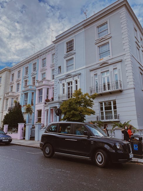 A street scene in a residential area featuring a row of tall, white Victorian-style terraced houses with decorative mouldings, bay windows, and small balconies. The buildings are painted in light pastel shades with some architectural details highlighted by the daylight. In the foreground, a black London-style taxi with a rounded shape and glossy finish is parked on the street. Next to the taxi, a woman wearing a mask and red jacket is standing near a blue recycling bin, with other pedestrians visible on the pavement. A tall, leafy tree partially obscures one of the houses, adding greenery to the scene. The sky above is partly cloudy with patches of blue visible, providing natural light that highlights the textures of the building facades, the vehicle, and the street environment. The image conveys a typical urban residential setting suitable for private waste collection or on-site rubbish clearance, consistent with independent rubbish removal services in the area.