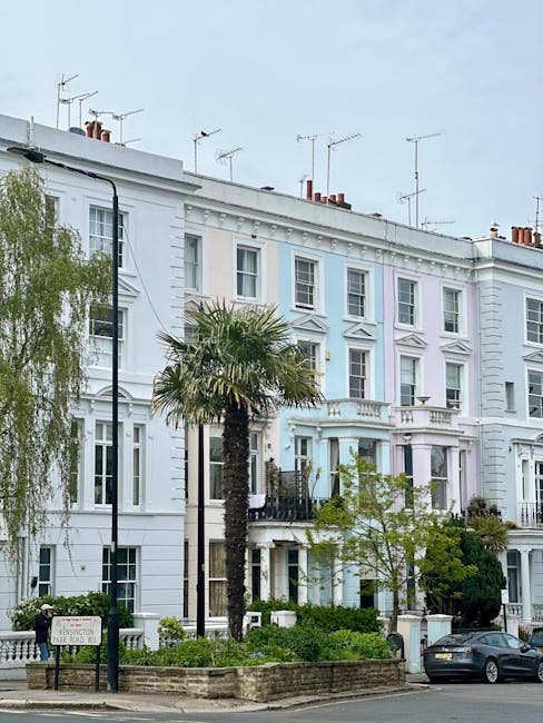 A street scene in a residential area featuring a row of tall, white Victorian-style terraced houses with decorative mouldings, bay windows, and small balconies. The buildings are painted in light pastel shades with some architectural details highlighted by the daylight. In the foreground, a black London-style taxi with a rounded shape and glossy finish is parked on the street. Next to the taxi, a woman wearing a mask and red jacket is standing near a blue recycling bin, with other pedestrians visible on the pavement. A tall, leafy tree partially obscures one of the houses, adding greenery to the scene. The sky above is partly cloudy with patches of blue visible, providing natural light that highlights the textures of the building facades, the vehicle, and the street environment. The image conveys a typical urban residential setting suitable for private waste collection or on-site rubbish clearance, consistent with independent rubbish removal services in the area.