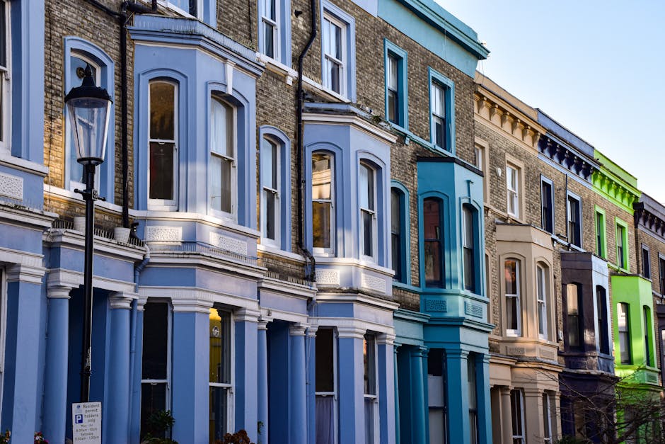 A row of multi-story terraced houses with Victorian-style architectural features, including large bay windows, decorative cornices, and brick facades. The buildings are painted in a variety of pastel and muted colors such as light blue, teal, beige, dark navy, and bright lime green, creating a vibrant and diverse streetscape. Each house has tall, narrow windows with white frames, some with curtains visible inside. A black street lamp is positioned in the foreground on the left side of the image, casting a subtle shadow on a paved sidewalk. The ground level of the buildings features decorated window surrounds and small front gardens or steps leading to front doors not visible in the photo. The sky above is clear with natural daylight illuminating the scene, emphasizing the textures of the bricks and the architectural details. This image depicts an urban residential area typical of cities with ornate private housing, which could be cleaned or cleared of waste by companies such as Waste Collection Notting Hill as part of their rubbish removal services, especially when preparing properties for renovation or disposal of household items.